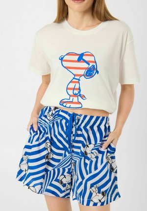 Femme portant un t-shirt blanc avec un imprimé Snoopy rayé orange et bleu et un short rayé bleu et blanc avec de petites figures de Snoopy.