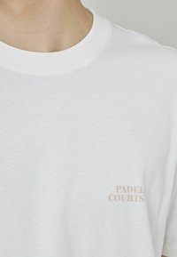 T-shirt en coton blanc avec une texture douce, col rond, et le texte "PADEL COURTS" imprimé en rose clair sur le côté gauche.