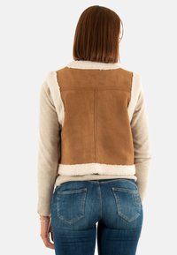 Brun fuskmockajacka med ett fårskinnsliknande foder längs kanterna, bärs över en beige tröja. Välpassade denimjeans runder av outfiten.