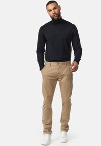 Jersey de cuello alto negro, chinos beige y zapatillas blancas. Los chinos son de corte recto con bolsillos delanteros y trabillas para cinturón. Textura de tela suave.