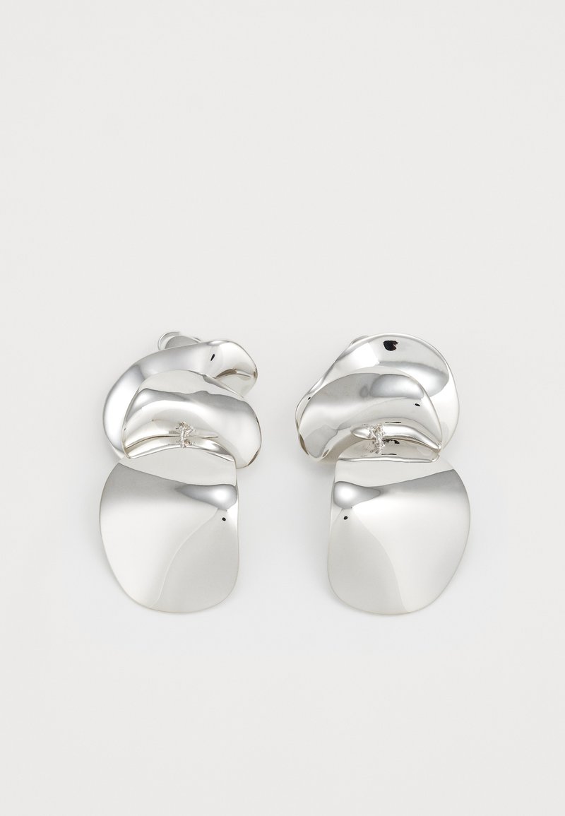 Ezüst fém fülbevalók, amelyek szobrászati, csavart formával rendelkeznek, sima és fényes felületekkel, kerek, lapos alakzatokkal kiegészítve.