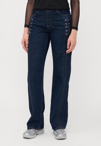 Mørkeblå denimjeans med rette ben, med dekorativ snøring langs sidesømmer og kontrasterende oransje sømdetaljer.