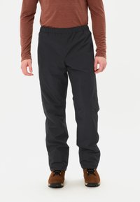 Pantaloni neri impermeabili con vita elasticizzata e vestibilità ampia, indossati sopra stivali da escursionismo marroni, abbinati a una maglietta marrone a maniche lunghe.