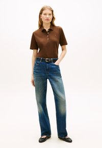 Γυναίκα με καφέ κοντομάνικο πόλο, ξεθωριασμένο μπλε τζιν με μαύρη ζώνη και μαύρα μπαλαρίνια, στέκεται με το ένα χέρι στην τσέπη.