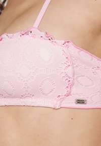 Bralette de renda rosa com um padrão circular, bordas em formato de onda e alças finas. Inclui uma pequena etiqueta "Banana Moon" na lateral.