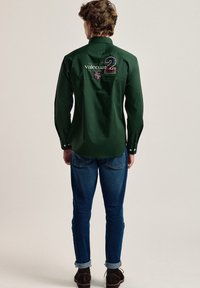Camisa verde de botones y de manga larga, con un logo y texto en la parte de atrás. Combinada con pantalones vaqueros azules y zapatos marrón oscuro.