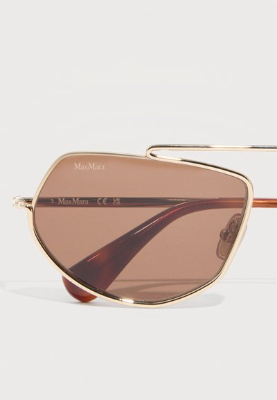 Ασύμμετρα γυαλιά ηλίου με χρυσή κορνίζα, καφέ φακούς με απόχρωση και λεπτό, κυρτό βραχίονα στο πλάι, εμφανή πάνω σε απλό φόντο.