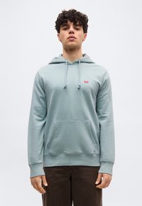 Νεαρός  με σγουρά μαλλιά που φοράει ανοιχτό μπλε φούτερ με κουκούλα, μπροστινή τσέπη και λογότυπο, στέκεται μπροστά σε μια απλή ανοιχτόχρωμη επιφάνεια.