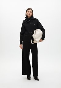 Prenda negra con un diseño estructurado, que presenta acentos y botones dorados, combinada con un bolso blanco suave. La modelo lleva unos elegantes tacones negros.