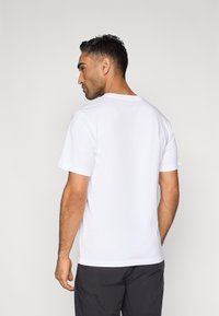 Άσπρο κοντομάνικο μπλουζάκι, στρογγυλή λαιμόκοψη, κατασκευασμένο από βαμβάκι, λεία υφή, πίσω όψη, φορεμένο με σκούρα σορτς. Χωρίς εμφανή σχέδια ή διακοσμητικά στοιχεία.