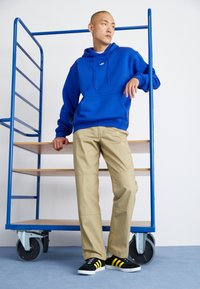 Sudadera con capucha azul, pantalones beige y zapatillas negras con rayas amarillas. Diseño simple, tejido suave, con un ajuste relajado y detalles de costura visibles.