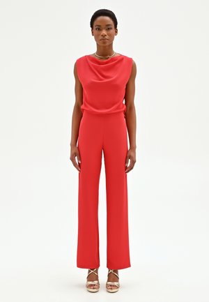 Donna in piedi di fronte, indossa una tuta rossa senza maniche e sandali dorati con cinturini, su uno sfondo bianco.