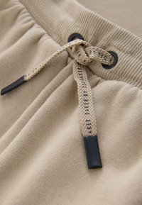 Gros plan sur la ceinture d'un pantalon de survêtement beige avec un cordon tressé noué, comportant un texte noir et des embouts en plastique noir.