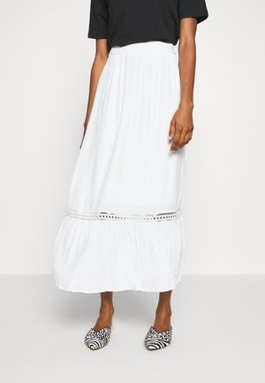 Femme portant une jupe midi blanche taille haute avec bordure en dentelle et des ballerines pointues à motifs noir et blanc.