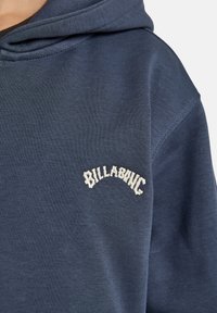 Granatowa bluza z kapturem wykonana z miękkiej bawełny miksowanej z teksturowanym wykończeniem. Posiada białe haftowane logo "BILLABONG" na lewej części klatki piersiowej.