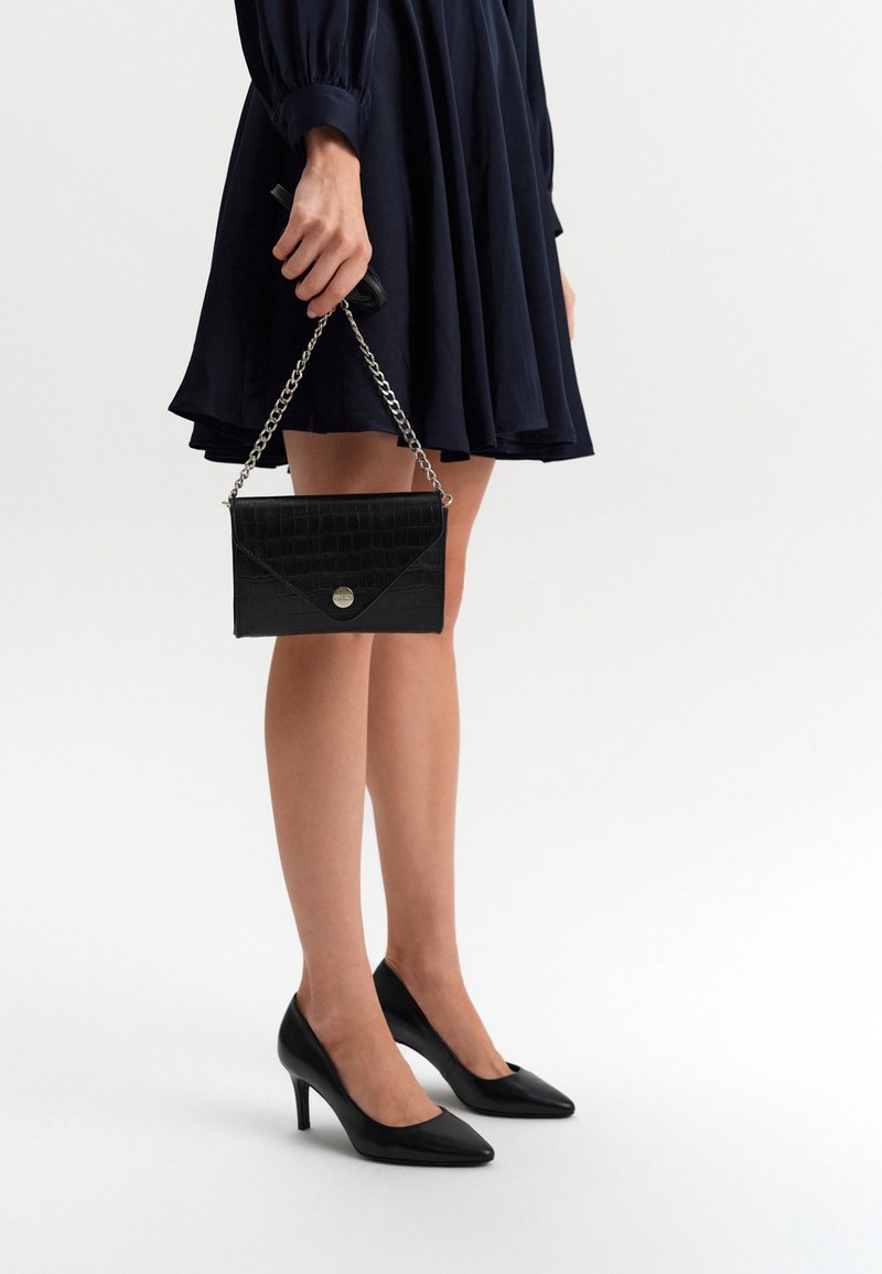 Czarna torebka kopertowa z tłoczonym wzorem w krokodyle, z srebrnym łańcuchem, trzymana w jednej ręce. Nosi ją w połączeniu z granatową sukienką i czarnymi szpilkami.
