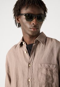 Camicia di lino marrone chiaro con taschino, modello con bottoni e una texture sottile. Indossata sopra una maglietta nera, abbinata a occhiali da sole scuri.