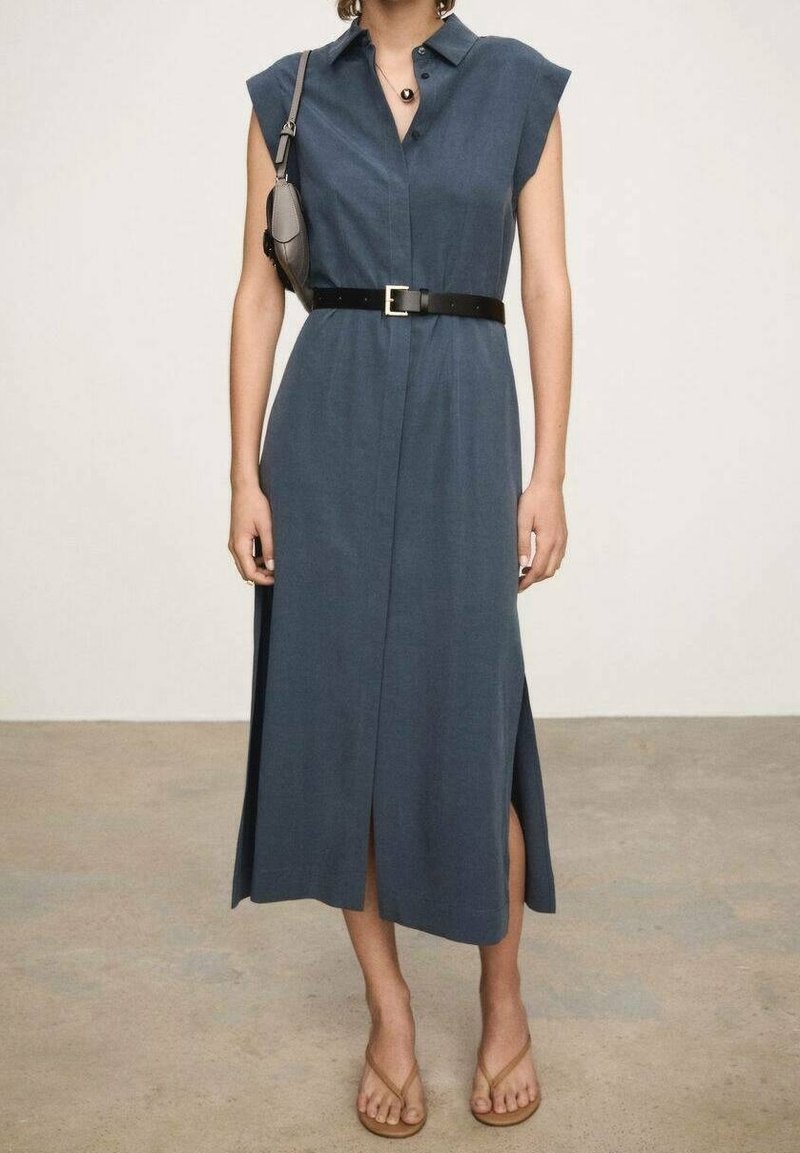 Robe midi bleue sans manches avec fentes latérales, ceinture noire, petit sac à bandoulière, et sandales beige sur une personne debout.