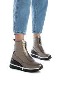 Botas tobilleras metálicas de bronce con tacón cuña, que cuentan con un zipper frontal y textura acolchada, combinadas con unos jeans cortos de color azul claro.