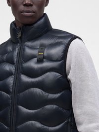 Homme portant une veste noire matelassée sans manches avec un col montant et une chemise à manches longues gris clair en dessous.