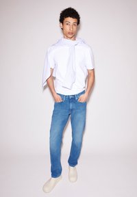 T-shirt blanc, jeans bleu clair, sabots crème ; tissu rayé drapé autour du cou ; le mannequin se tient avec les mains dans les poches devant un fond clair.