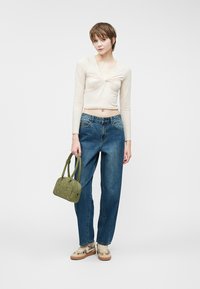 Top beige de manga larga con detalle retorcido, combinado con unos jeans de talle alto azules. La modelo sostiene un bolso verde y lleva zapatillas beige con acentos.
