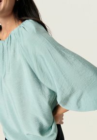Blusa leggera di colore azzurro chiaro con colletto rotondo, tessuto arricciato e maniche ampie e morbide. Materiale testurizzato e morbido che cade elegantemente.