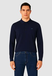 La polo shirt a maniche lunghe di colore blu navy presenta un colletto, tre bottoni frontali e un piccolo logo turchese sul lato sinistro del petto. Indossata con un jeans.