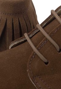 Scarpe in suede marrone con un design frangiato, caratterizzate da lacci e cuciture marroni chiari. La texture è morbida e opaca.