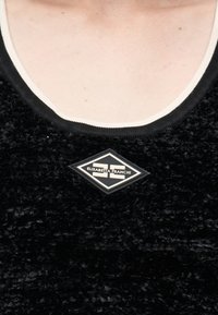 Κοντινή λήψη ενός ατόμου που φοράει μαύρο μπλουζάκι με υφή, με λευκή επένδυση στη λαιμόκοψη και κονκάρδα με το διαμάντι-σχήμα λογότυπο Elisabetta Franchi.