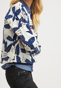 Giubbotto bomber a motivo floreale con foglie blu e fiori bianchi, con orlo e polsini a coste, indossato sopra una camicia di denim. Pantaloni neri visibili.