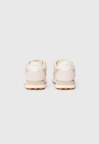 Light beige sneakers with suede and mesh panels, rounded shape, cushioned sole, and logo detailing on the heel. Flat lacing system.