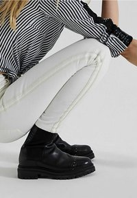 Botas de cuero negras hasta el tobillo con suelas acanaladas y adornos de tachuelas, combinadas con pantalones blancos ajustados que presentan costuras verticales y una blusa a rayas en blanco y negro.