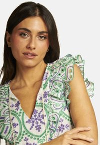 Blusa blanca con bordado floral en verde y púrpura, mangas con volantes y un escote en V profundo, con acentos en forma de lazo y detalle de cordón.