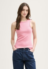 Różowy bezrękawnik z prążkowanej tkaniny o okrągłym dekolcie, w połączeniu z luźnymi, ciemnoniebieskimi dżinsami. Model ma ręce w kieszeniach.