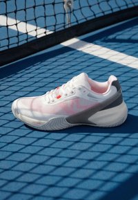 Chaussure de tennis blanche et grise sur un court bleu près d'un filet noir projetant des ombres sur la surface.