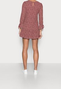 Frau in einem kastanienbraunen, floralen Mini-Kleid mit langen Ärmeln und weißen Sneakers, die vor einem schlichten weißen Hintergrund steht.