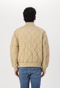 Chaqueta bomber acolchada beige con cuello y puños de canalé, que presenta un patrón de diamantes. Lleva puesta sobre unos jeans de color azul claro.