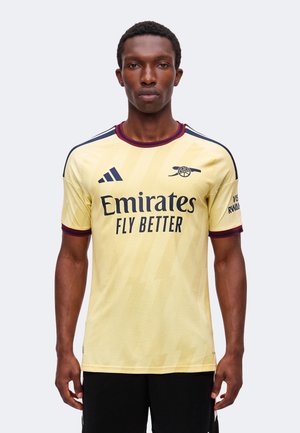 Modello maschile che indossa la maglia da calcio gialla dell'Arsenal con il logo "Emirates Fly Better" e lo stemma del club sul davanti, in piedi su uno sfondo semplice.