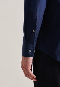 Marineblauwe overhemd met lange mouwen; heeft een knoopsluiting bij de manchetten en een subtiel logo op de mouw. Gladde textuur en strak ontwerp.