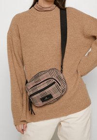 Pieni, tikattu olkalaukku, jossa on vaaleanpunainen ja musta ruutukuosi, vetoketjulliset osastot, musta hihna ja suorakulmainen logotunniste.