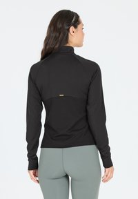 Schwarzes langärmliges Sportoberteil mit hohem Kragen, das im unteren Bereich eine gerippte Textur aufweist und ein kleines goldfarbenes Logo auf der Rückseite hat.