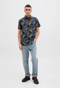 Camisa de botão de manga curta em tecido escuro com um padrão de flores e folhas em verde, azul e laranja; combinada com jeans azuis desbotados.