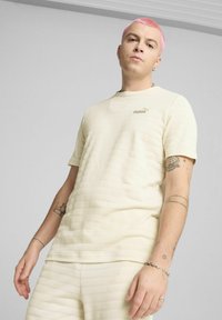 Cremefarvet tekstureret t-shirt med horisontale striber, der har et lille guld logo på brystet. Korte ærmer og rund halsudskæring.