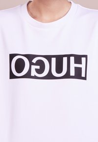 Osoba v bílém mikině s nápisem "HUGO" vytištěným černými obrácenými písmeny na hrudi.