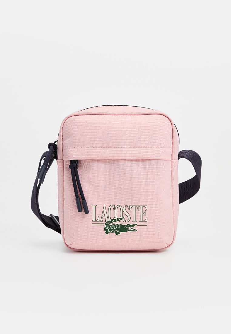 Sac à bandoulière en tissu rose avec fermeture éclair, présentant un logo Lacoste vert et des sangles d'accent en bleu foncé. Format carré compact.