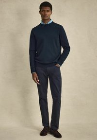 Marinblå crew-neck tröja över en ljusblå skjorta med krage, matchat med mörka byxor och bruna skor, med en mjuk tygtextur.