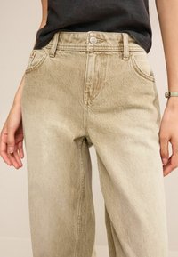 Jean beige taille haute en denim, avec fermeture à bouton, cinq poches et une coupe droite avec une texture subtile.