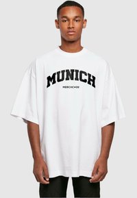 Bílá oversized tričko s tučným černým nápisem "MUNICH" a "MERCHC•DE" pod ním. Krátké rukávy, volný střih a měkký bavlněný materiál.