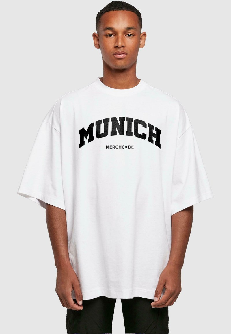 Bílá oversized tričko s tučným černým nápisem "MUNICH" a "MERCHC•DE" pod ním. Krátké rukávy, volný střih a měkký bavlněný materiál.